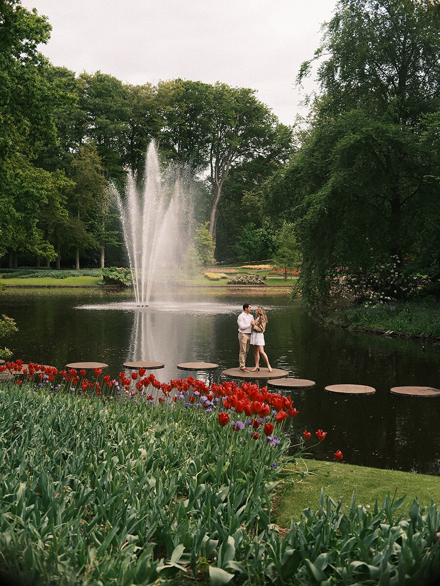 On stepping stones across a pond at Keukenhof, a couple stands close with a tall fountain behind and red tulips on the bank
