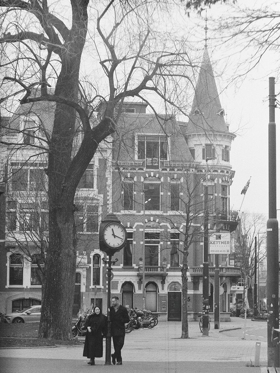 A street clock and turreted brick building tower above a couple in this wide black-and-white film portrait
