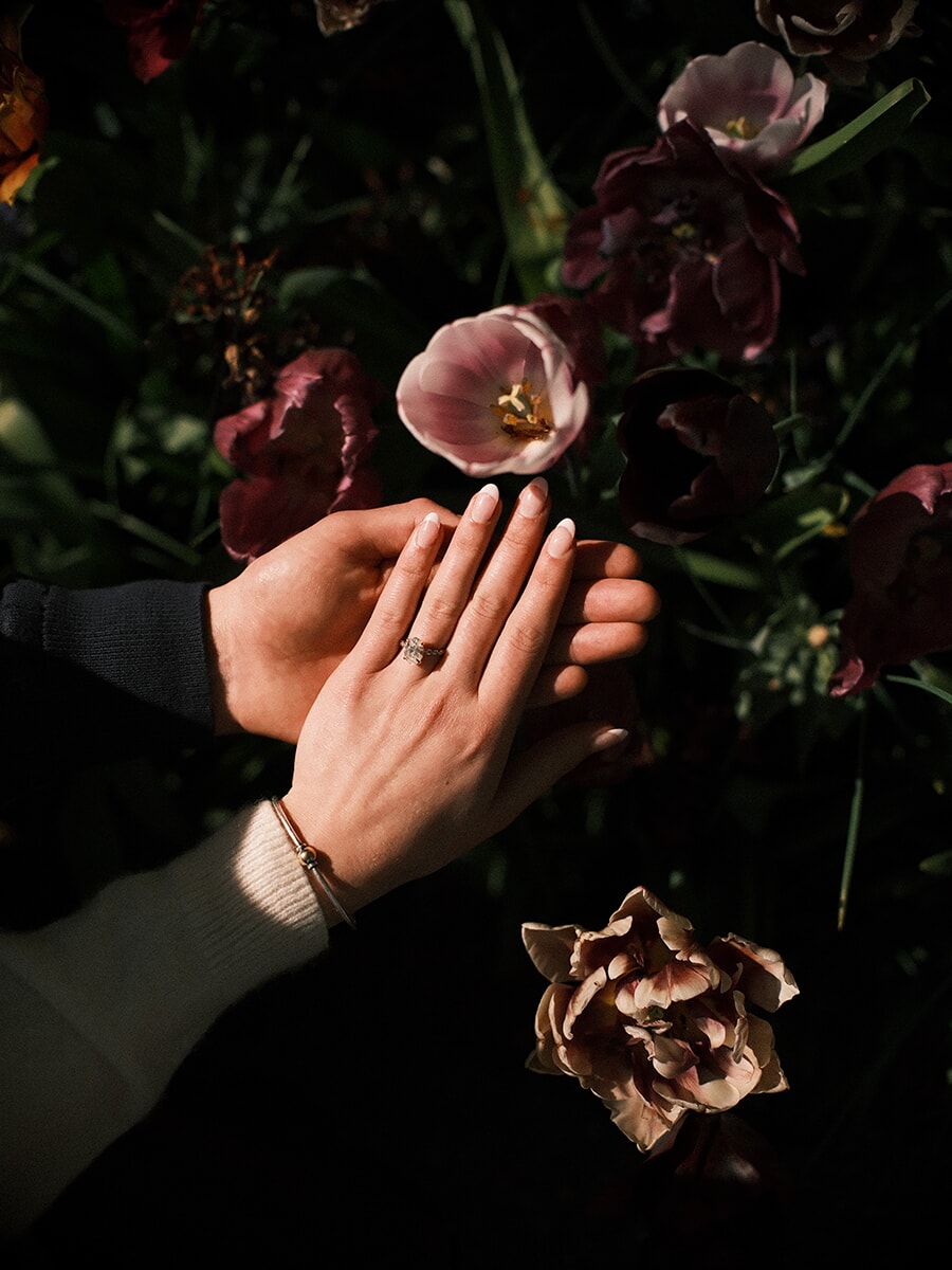 Engagement ring on her hand resting on his, surrounded by dark purple and pale pink tulips in low light