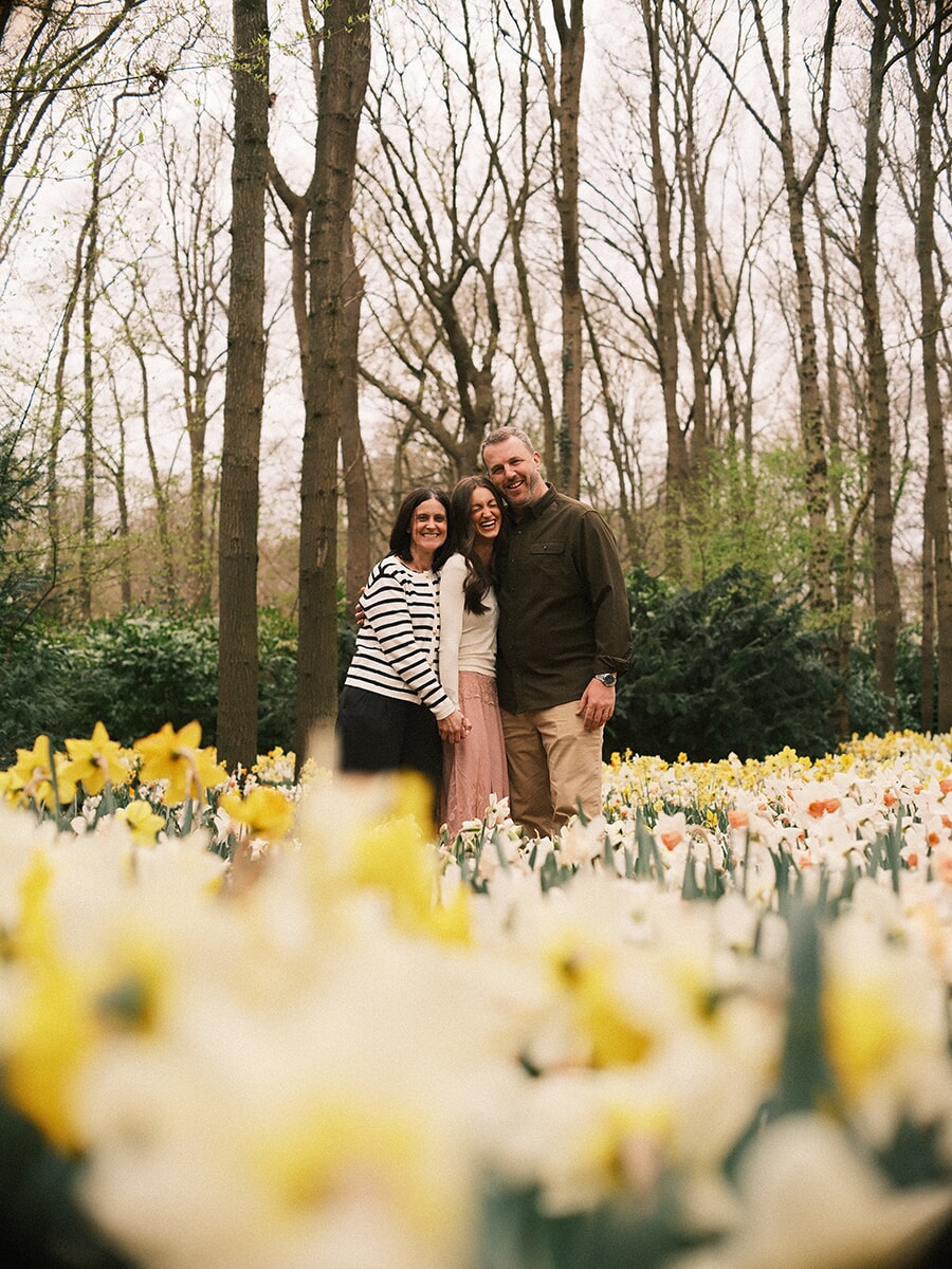 Three adults of one family laughing together among tall bare trees, white and yellow daffodils softly blurred in front