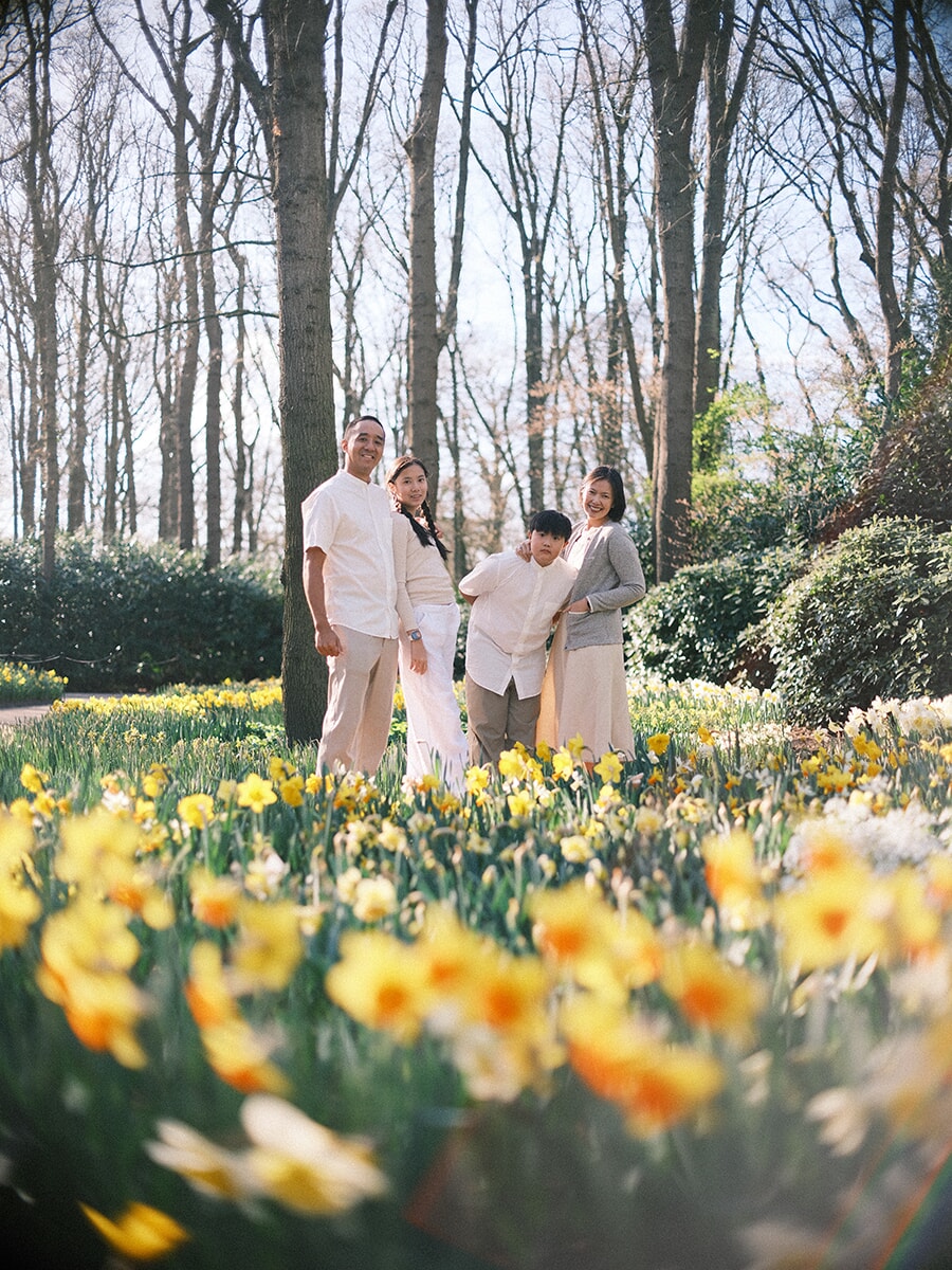 Family of four standing close among tall trees, yellow daffodils blurred in front during their family photoshoot at Keukenhof