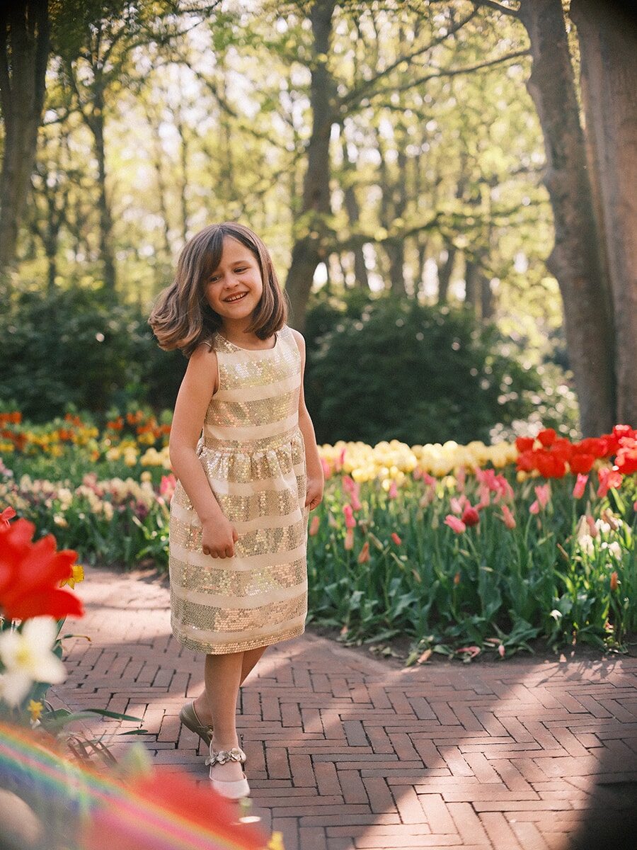 Little girl in a gold striped dress twirling on a herringbone brick path, tulip beds glowing behind her at Keukenhof