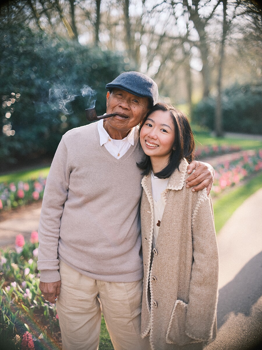 Grandfather in a flat cap with a pipe, his arm around his smiling granddaughter beside the tulip beds of Keukenhof