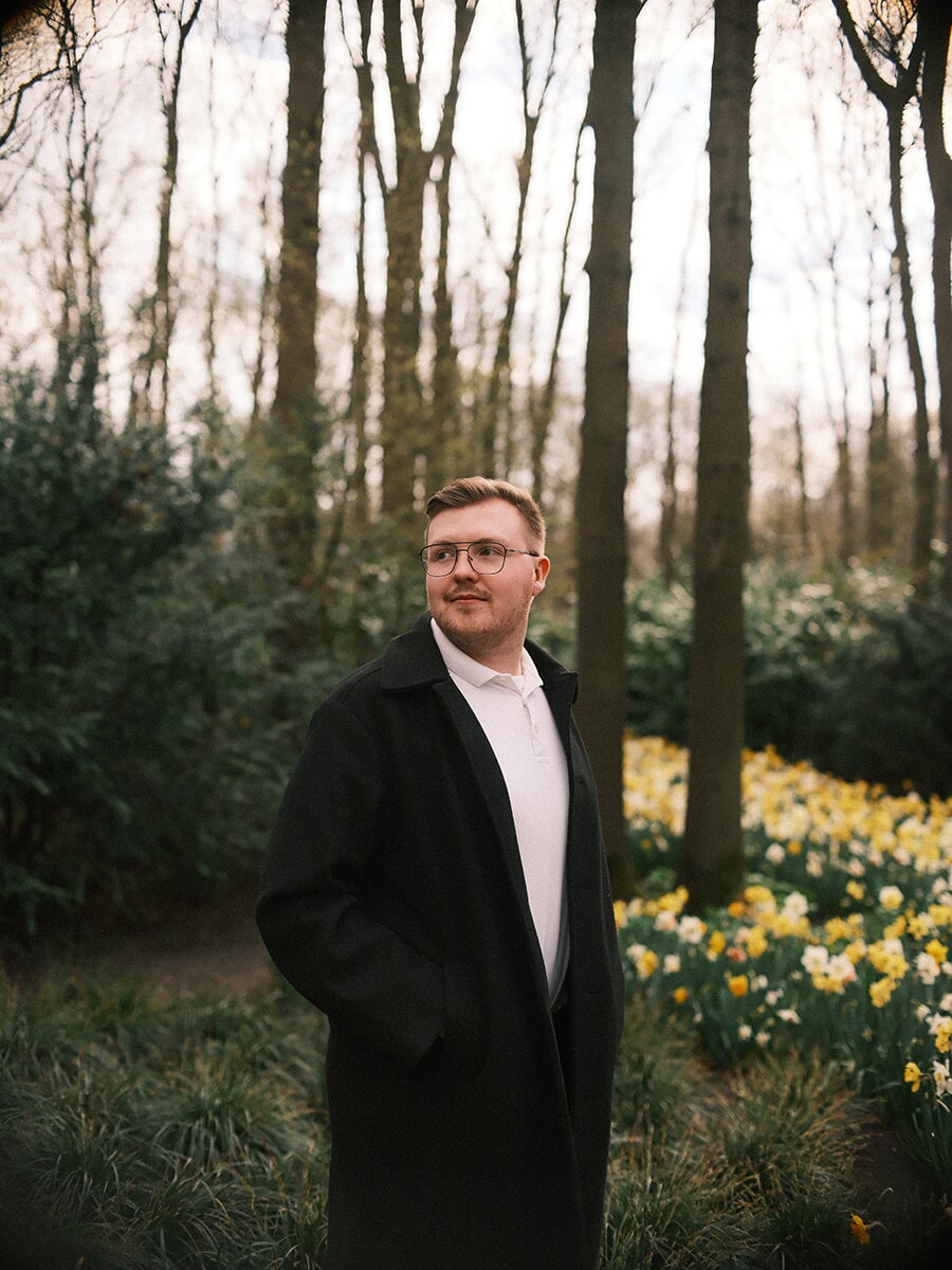 Man with glasses in a long black coat on a woodland path, drifts of yellow and white daffodils glowing beside him