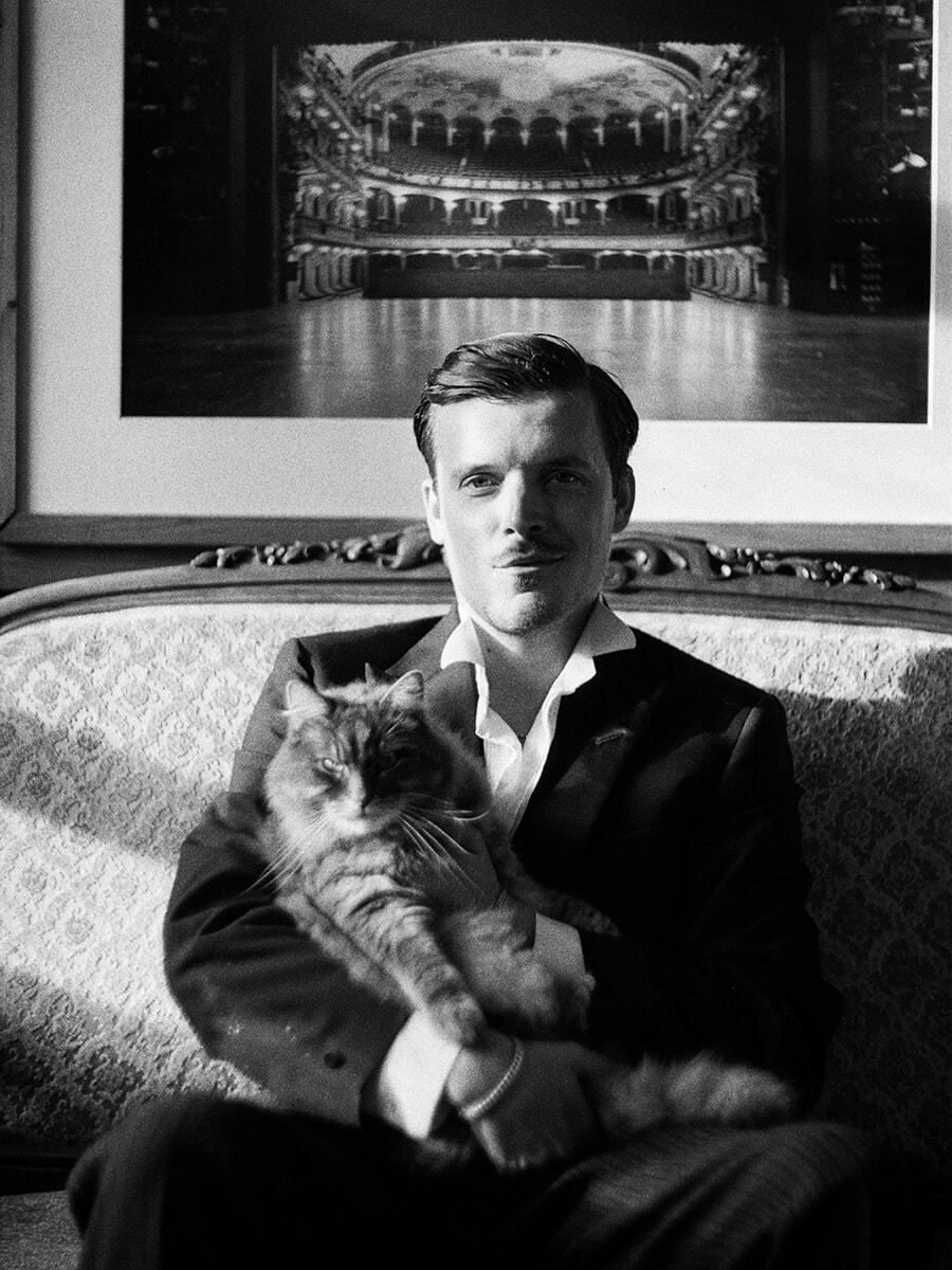 Seated on an ornate sofa, a man in a suit holds a cat on his lap in a black-and-white film portrait