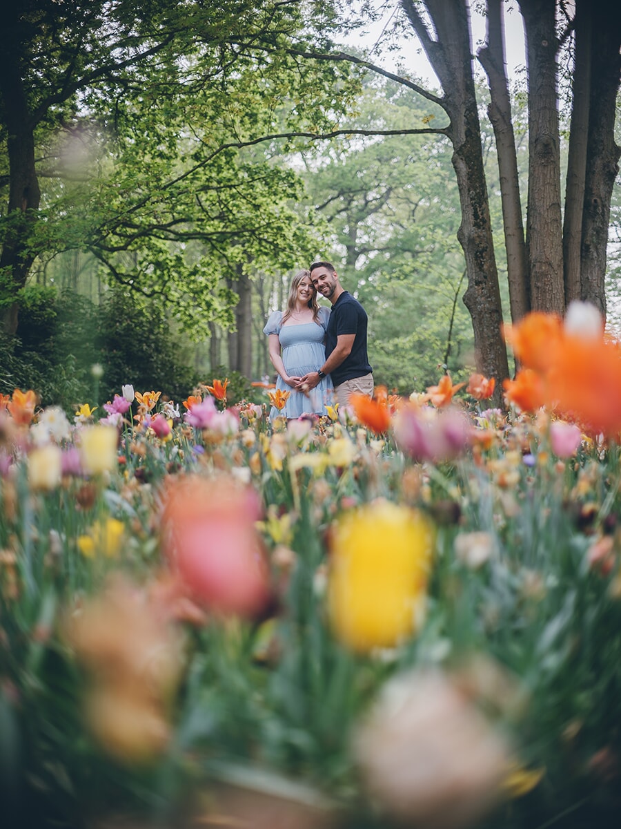 Expecting couple far back beneath green trees, large blurred tulips in pink, yellow and orange filling the foreground at Keukenhof