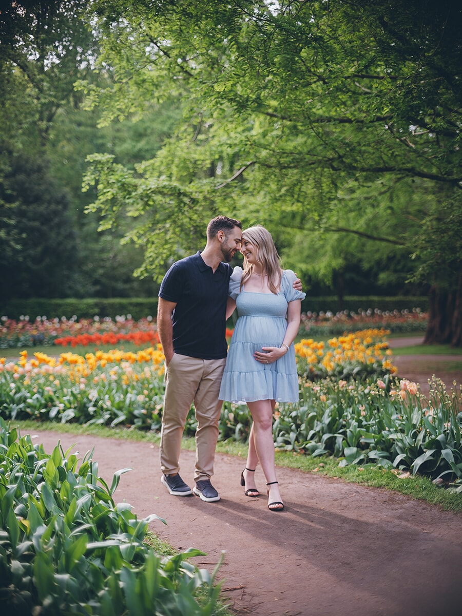 Expecting couple walking a garden path with foreheads close, her hand on her bump, during a maternity photoshoot at Keukenhof