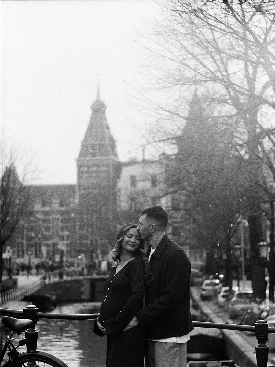 Analog maternity portrait of a couple sharing a forehead kiss on an Amsterdam canal bridge, with the Rijksmuseum behind them