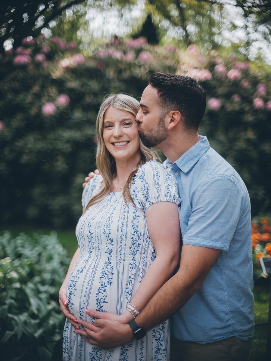 He kisses her temple as she smiles and cradles her bump in a blue floral dress, pink rhododendrons behind