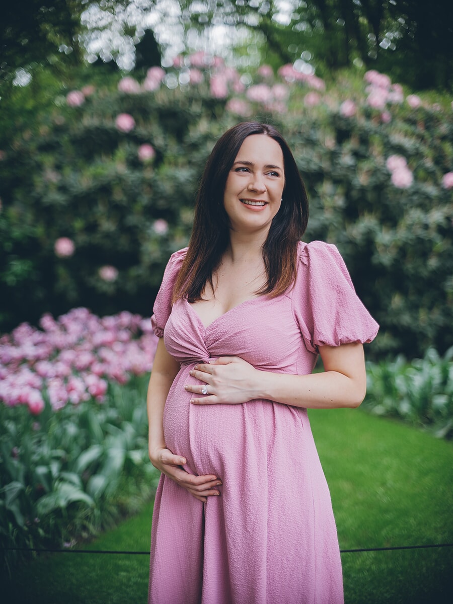 Mother-to-be in a dusty pink dress cradling her bump, smiling beside pink tulips and rhododendrons