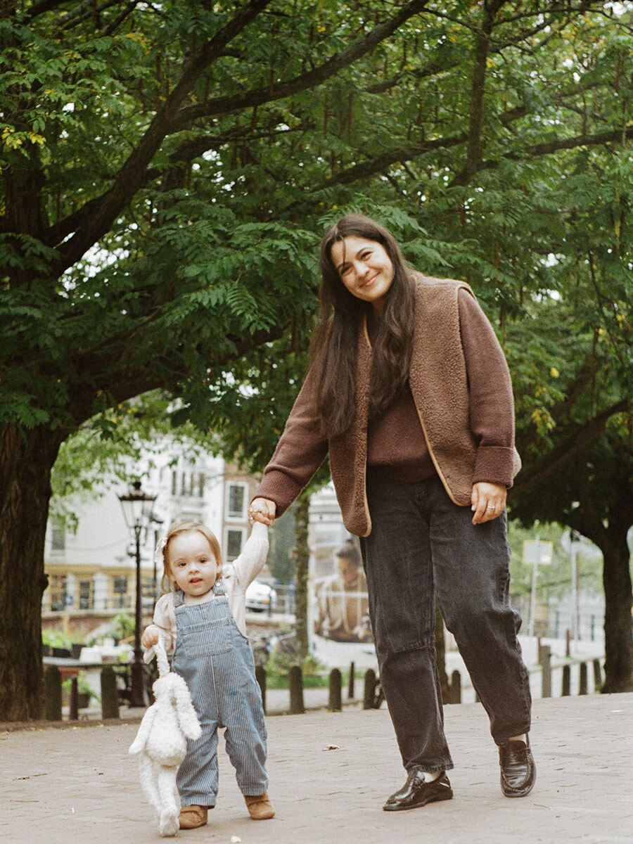Analog family portrait in Amsterdam of a mother holding her toddler’s hand beneath leafy trees, the child carrying a white rabbit toy