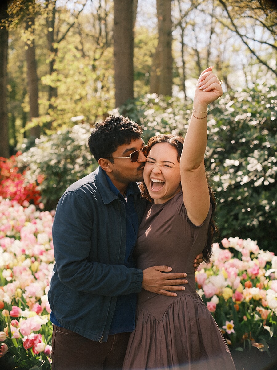 Newly engaged woman laughing with her fist raised as he kisses her cheek among white and pink tulips at Keukenhof