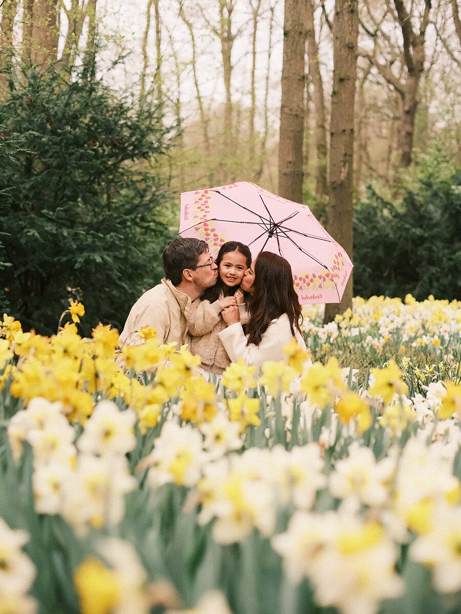 Parents kissing their little daughter's cheeks under a pink umbrella, sitting among yellow and white daffodils