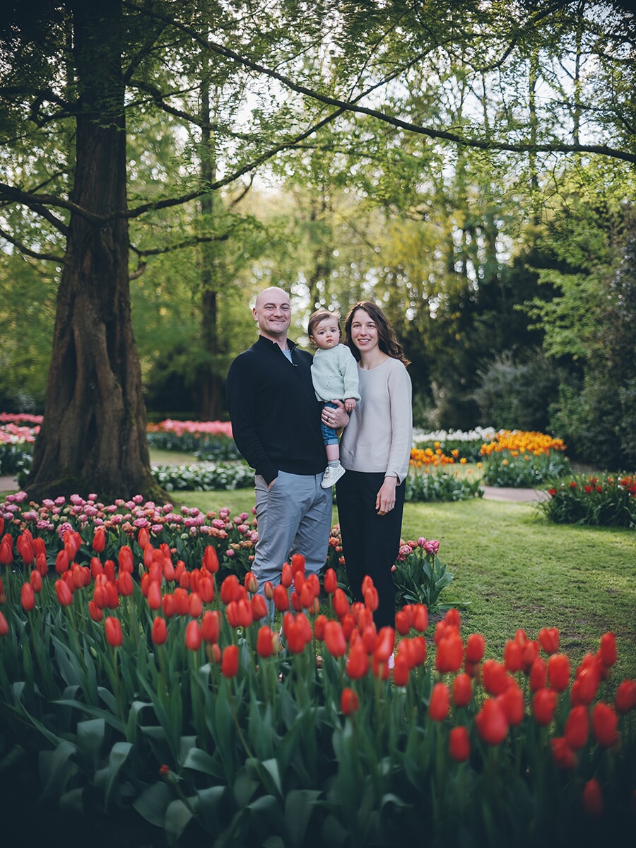 Parents holding their toddler under a big tree, red tulips in front and colorful beds across the lawn at Keukenhof