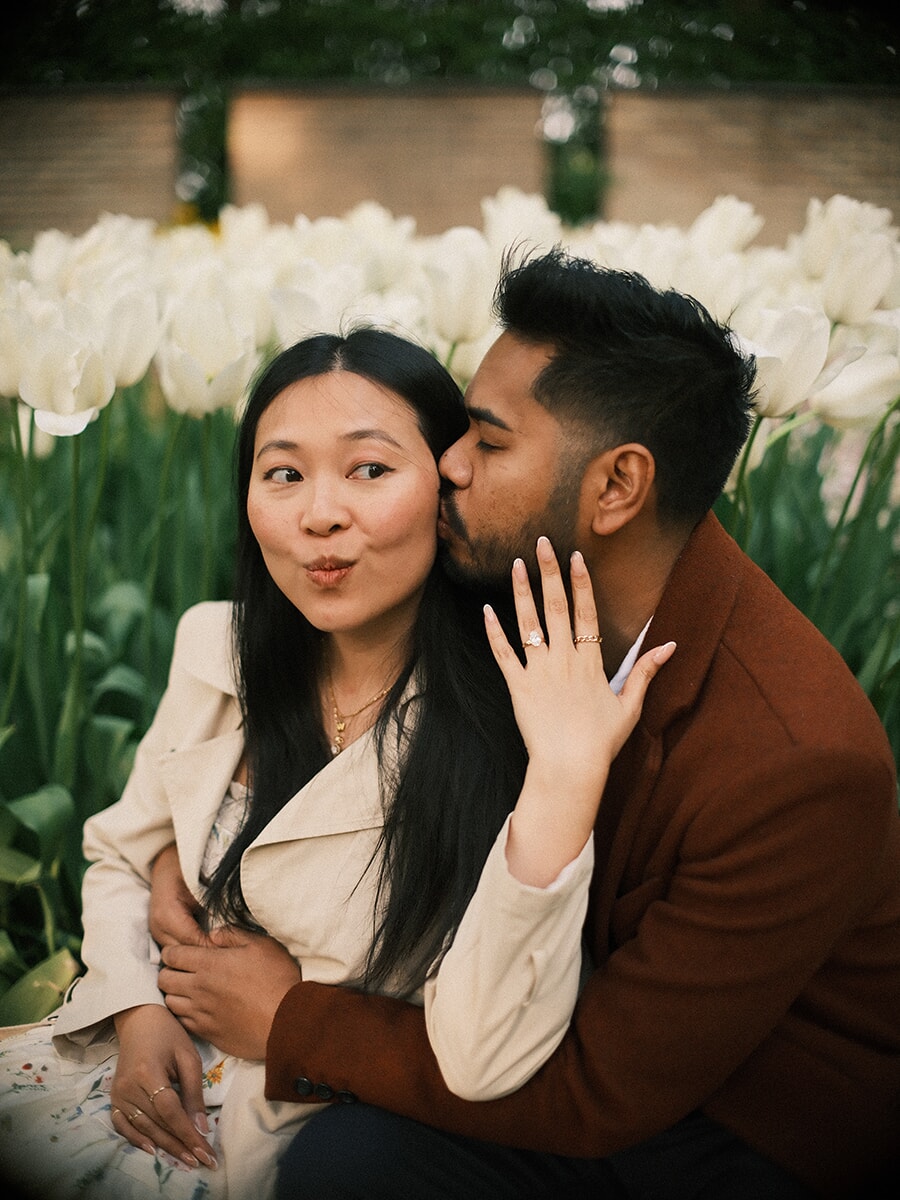 He kisses her cheek as she pouts playfully and shows her engagement ring, tall white tulips behind them
