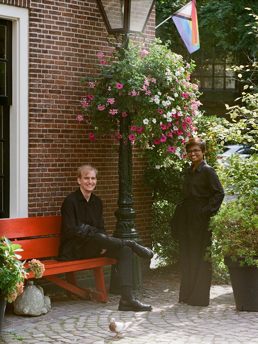 Two people in black smile at the camera by a red bench and hanging flowers, one seated and one standing