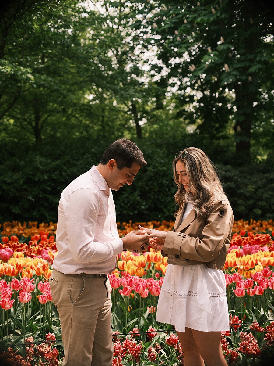 Man slipping an engagement ring onto her finger as she laughs, a surprise proposal among the tulip beds of Keukenhof