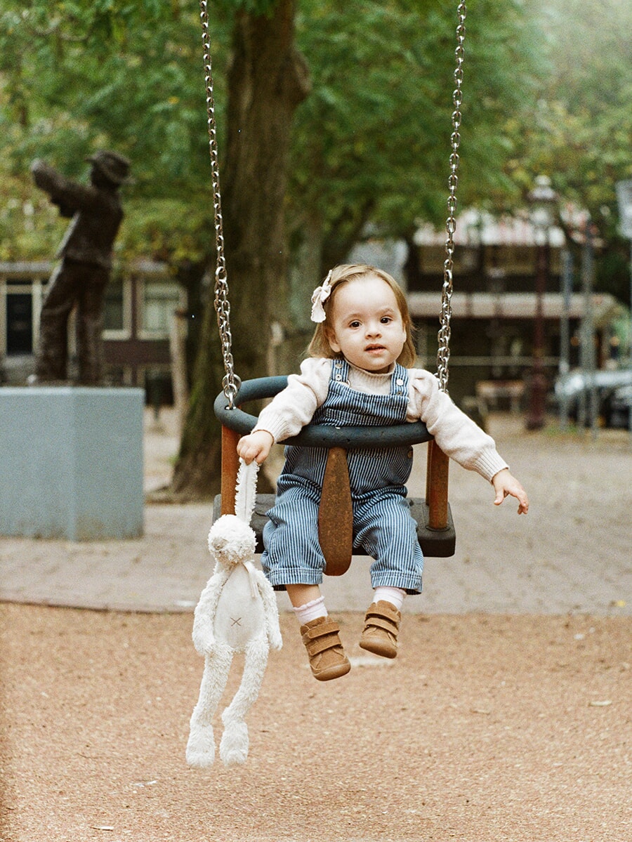 A toddler in blue striped dungarees sits in a playground swing holding a white rabbit toy, captured on colour film
