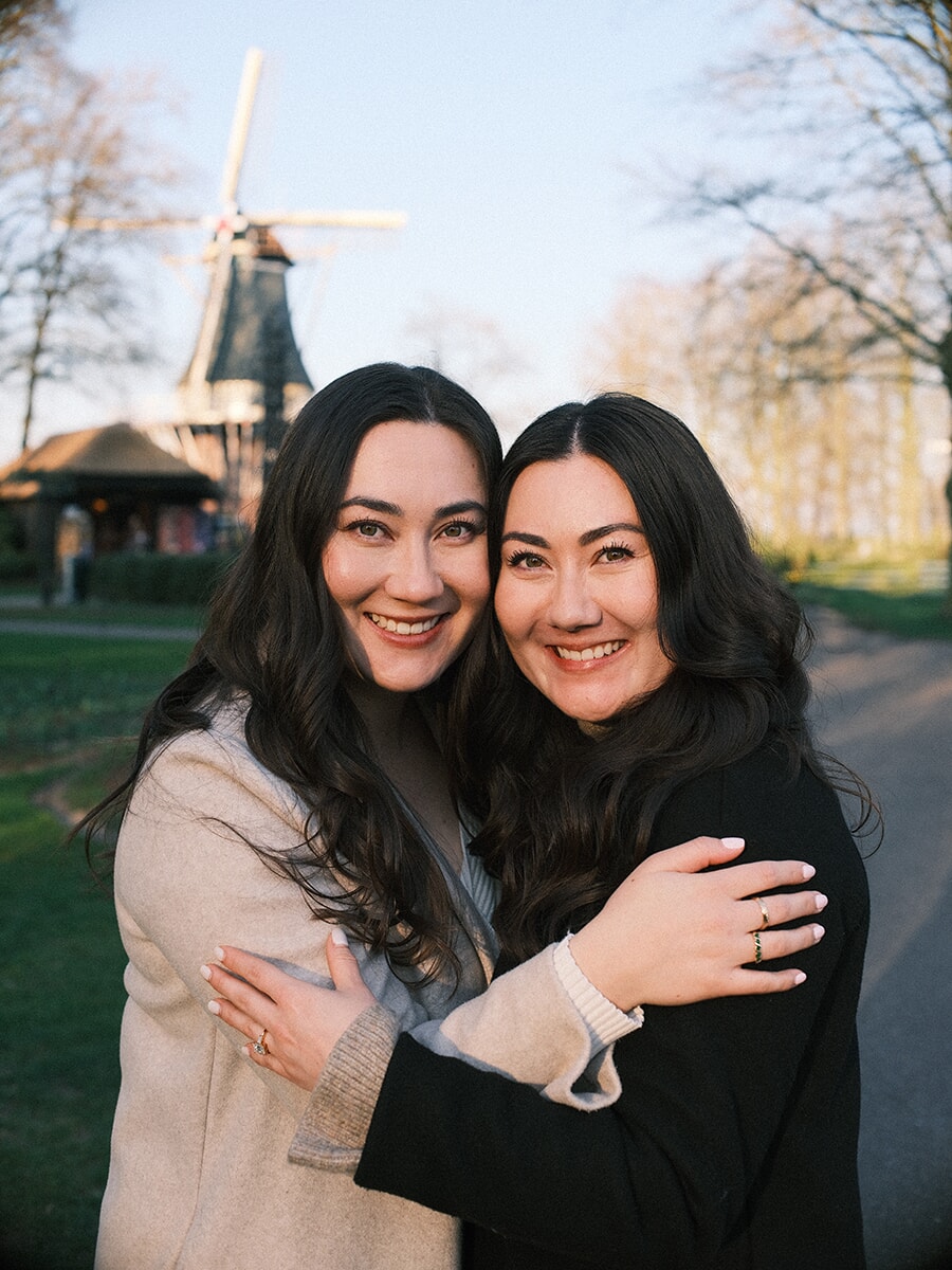 Two women hugging cheek to cheek in evening light, the historic windmill of Keukenhof rising behind them