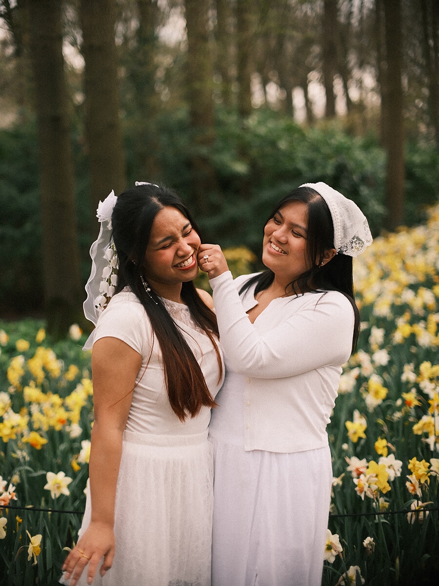 Two young women in white dresses and lace veils laughing as one pinches the other's cheek, daffodils behind