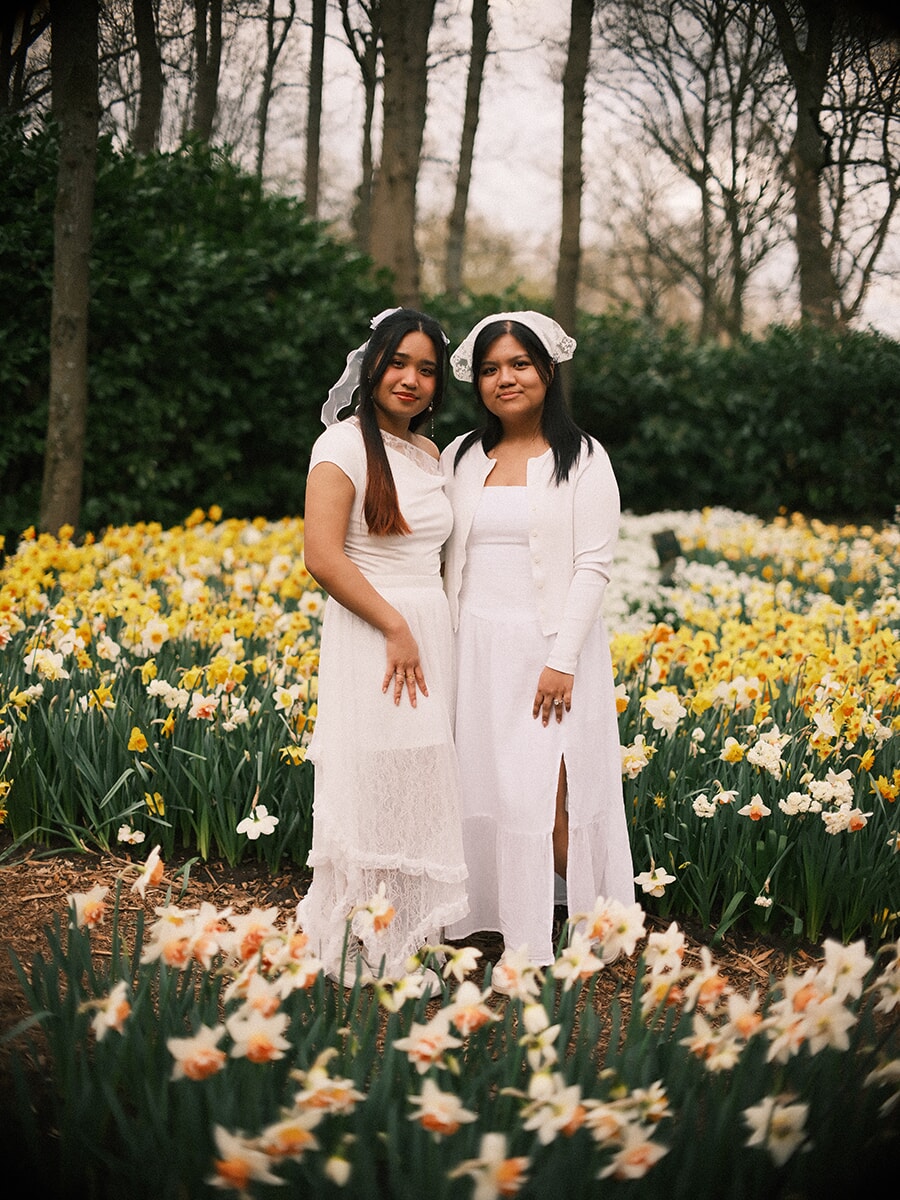 Two young women in white dresses and lace headpieces standing side by side in a daffodil bed at Keukenhof