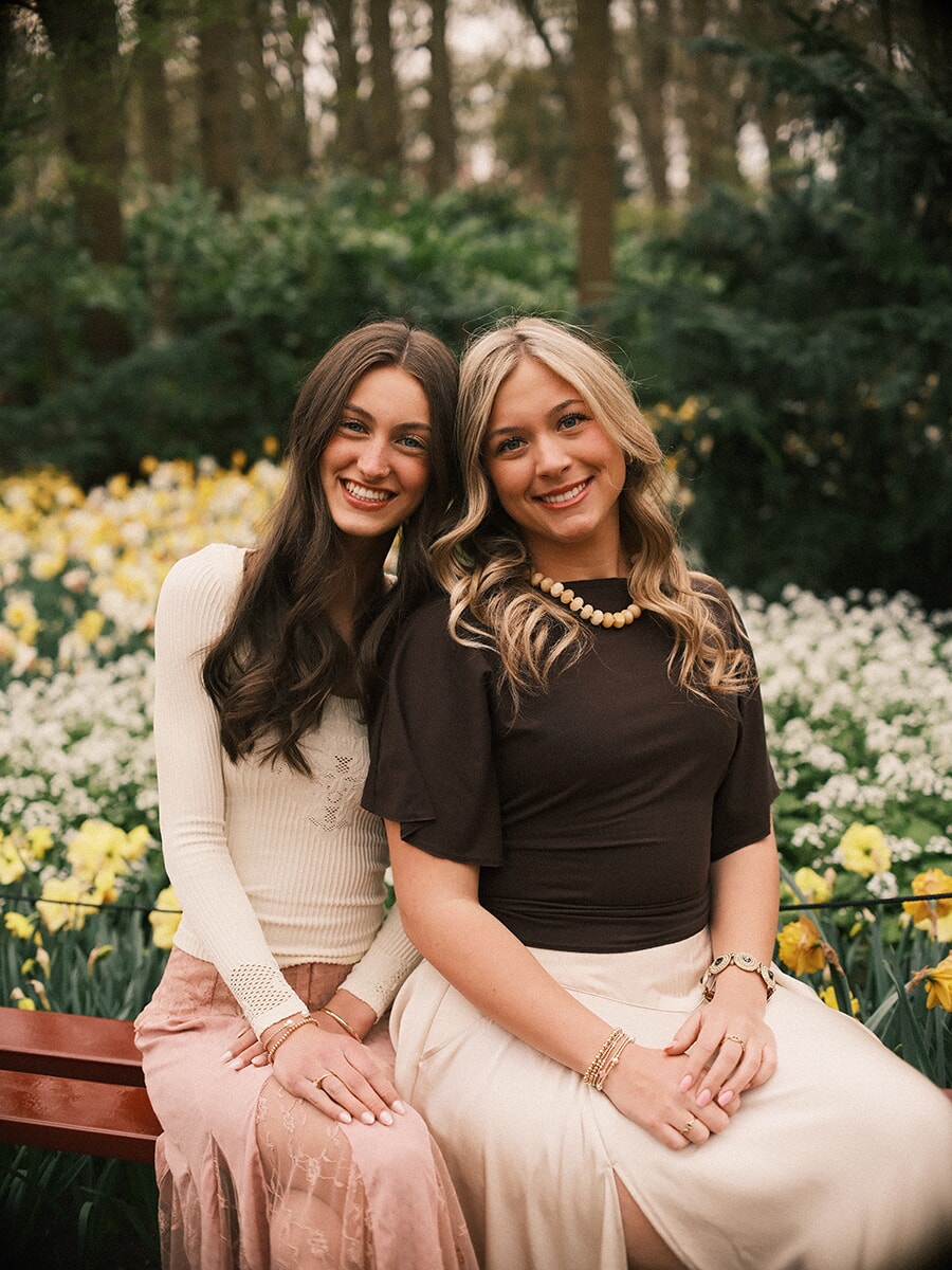 Two young women sitting close on a wooden bench, smiling in front of white and yellow daffodils