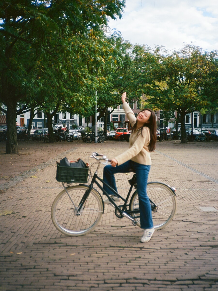 Raising one arm above her bicycle, a smiling woman in a cream cardigan poses on a brick square beneath leafy trees