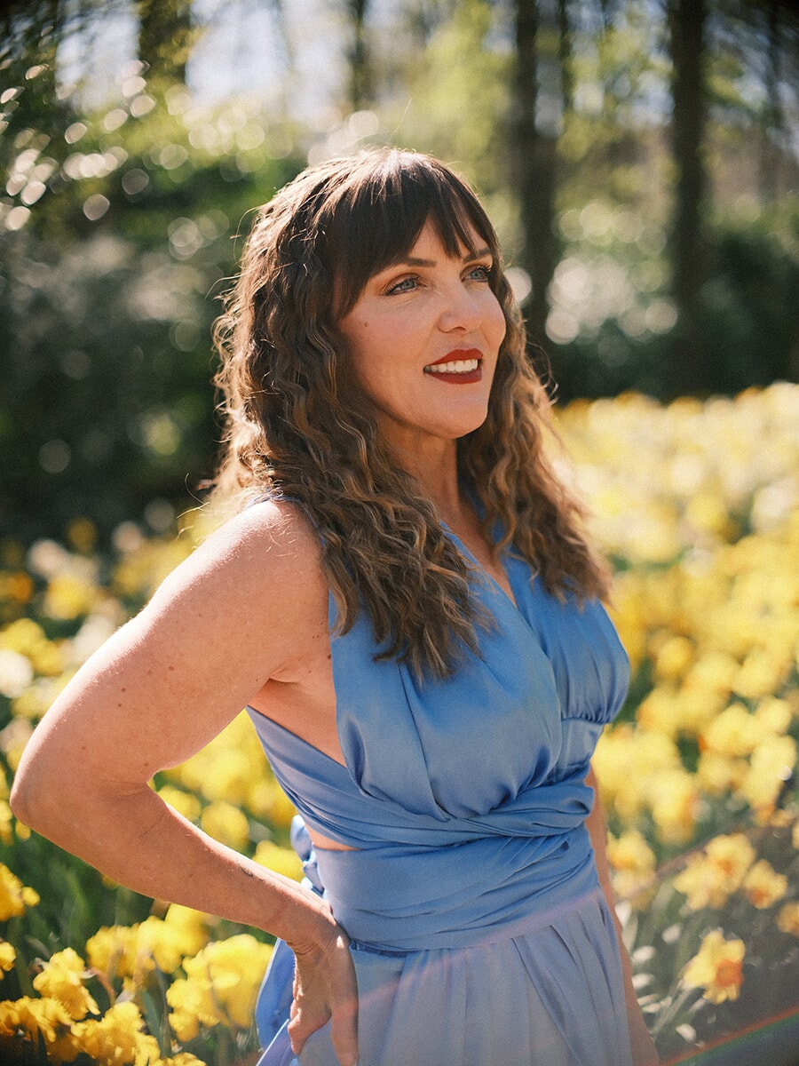 Woman with wavy hair and red lipstick in a sky-blue dress, smiling against sunlit daffodils and sparkling bokeh
