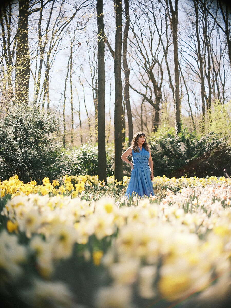 Standing in a field of daffodils under tall bare trees, a woman in a blue gown during her portrait session at Keukenhof