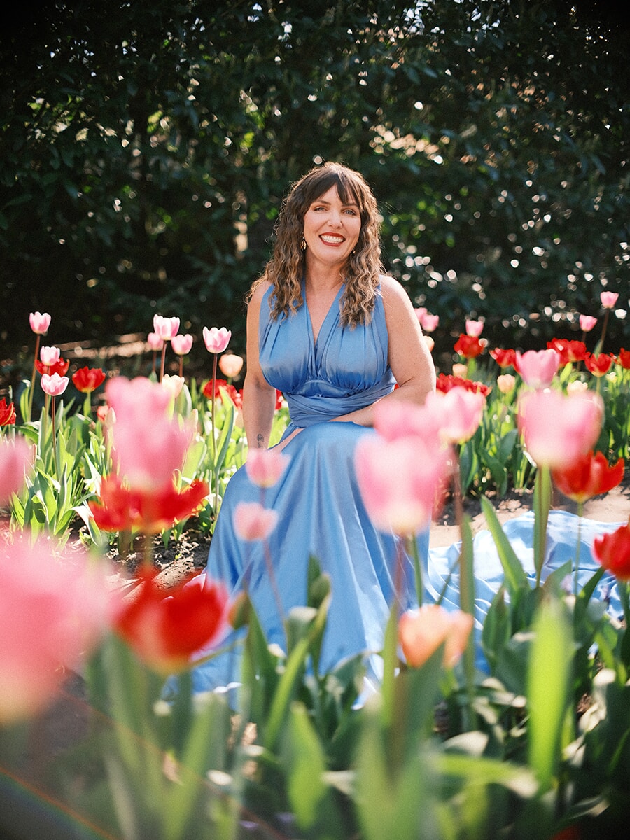 Laughing woman seated among pink and red tulips, her blue gown spread around her and blooms blurred in front