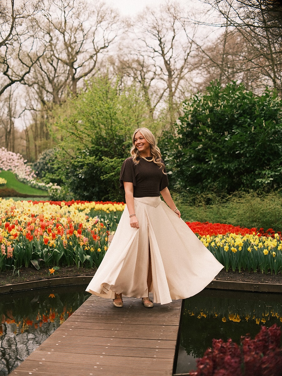Woman in a swirling cream skirt crossing a wooden boardwalk between two ponds, red and yellow tulips behind her at Keukenhof