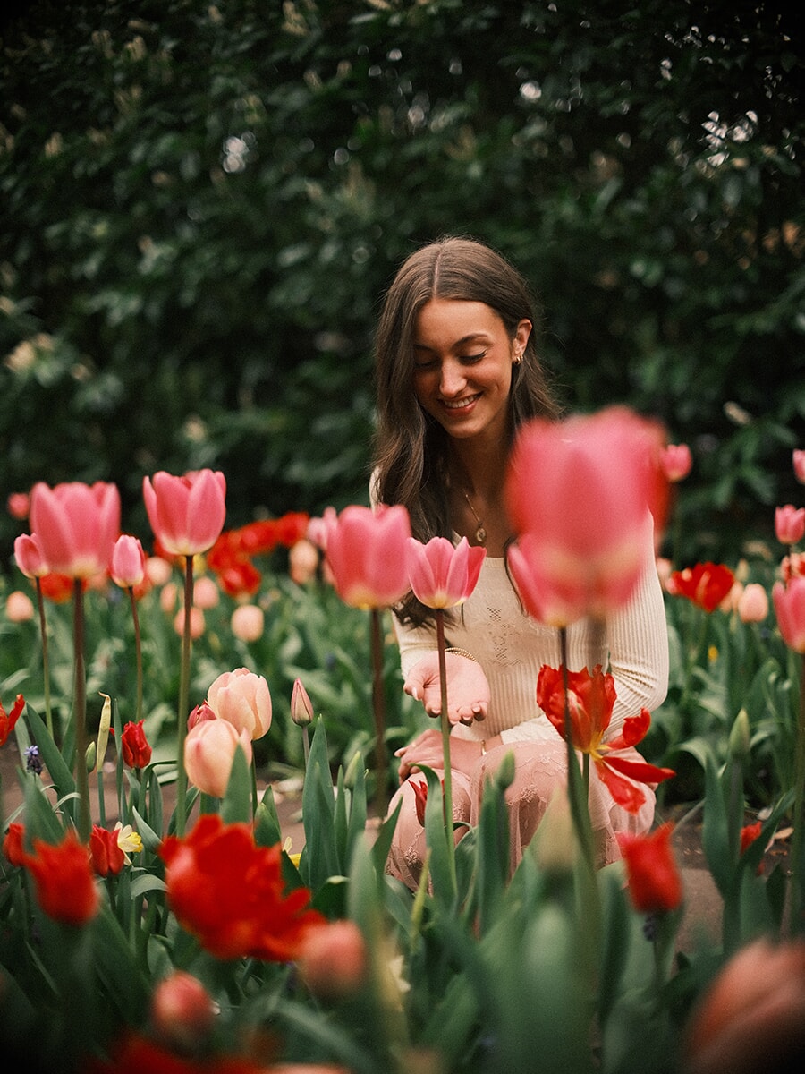 Smiling woman crouching low among pink and red tulips, soft blooms blurred in the foreground of a spring portrait