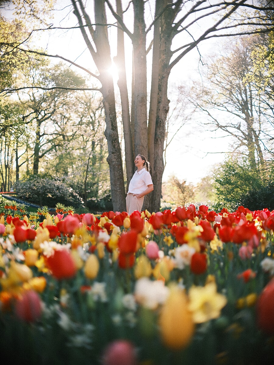 Woman in a white shirt laughing against a tree as the sun flares through the branches, red and yellow tulips in front