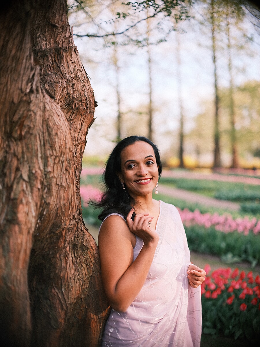 Smiling woman leaning against a tree trunk, pink and red tulips blurred behind her in a relaxed portrait at Keukenhof