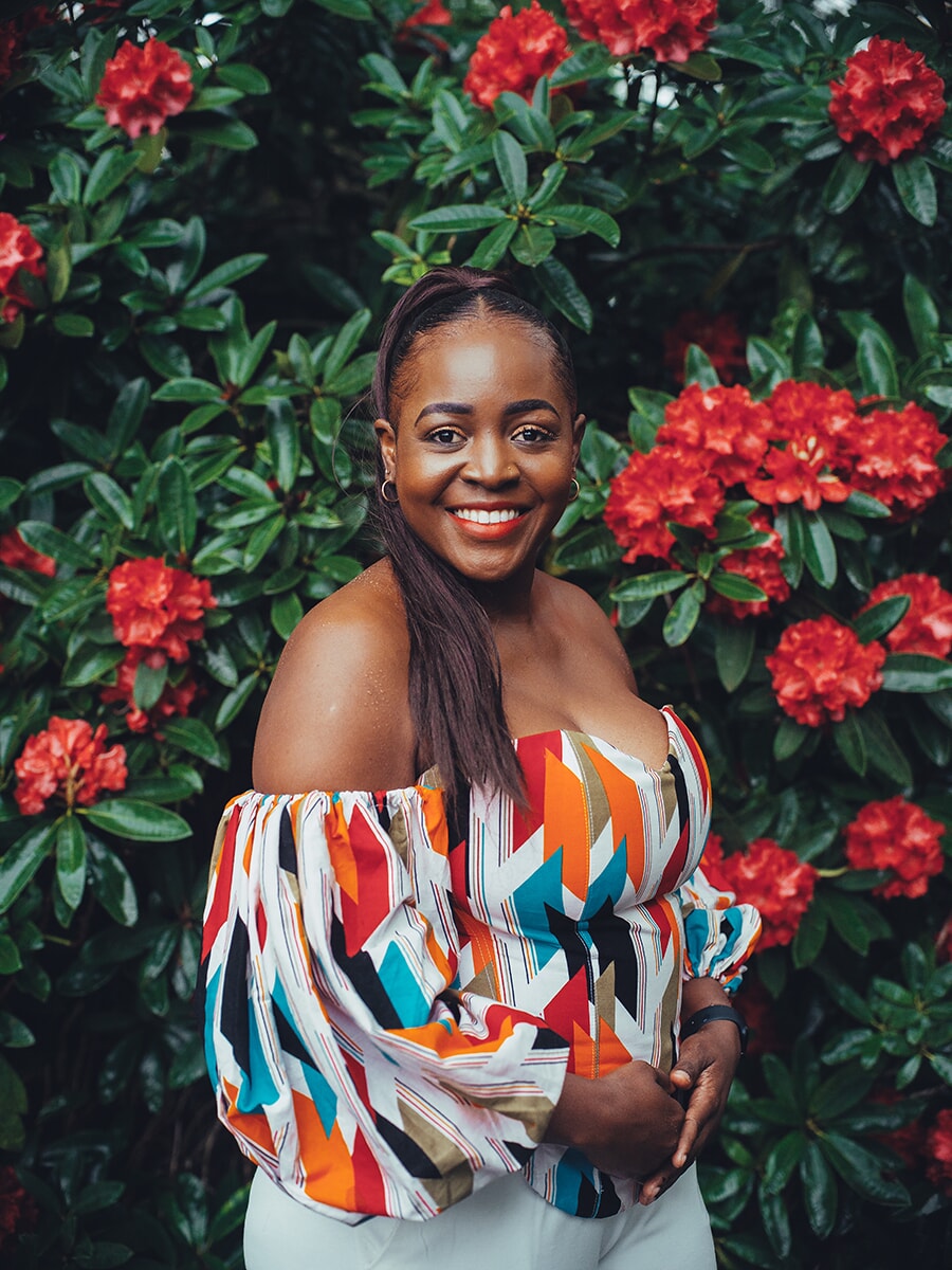 Woman with long braids in a colorful off-shoulder top, beaming in front of red rhododendron blooms, a vibrant spring portrait