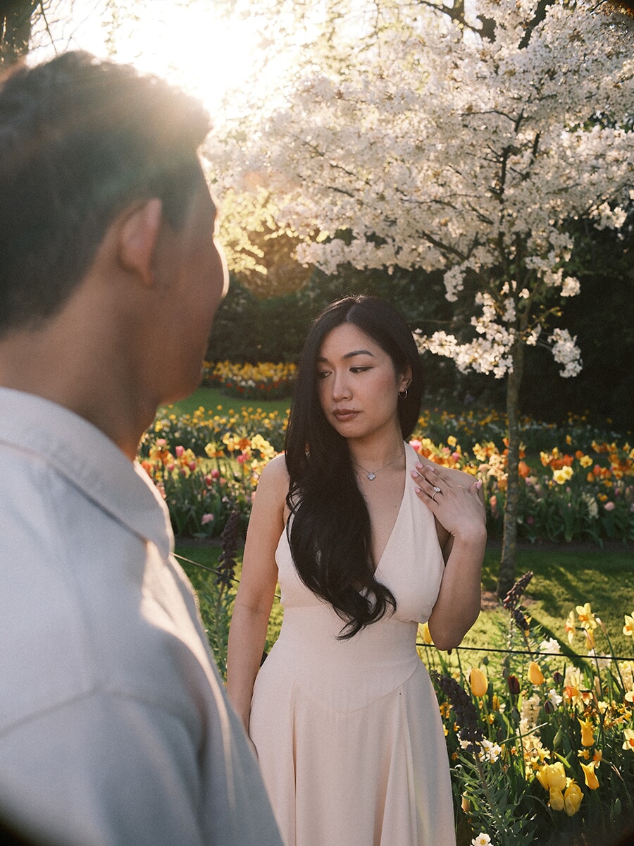Seen over his shoulder, she looks down in a peach halter dress beneath white cherry blossom during their couple photoshoot at Keukenhof