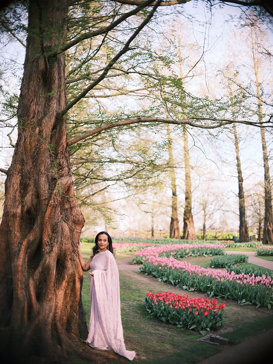 Woman in a pale pink draped dress beside a huge old tree trunk, curving beds of pink and red tulips behind her