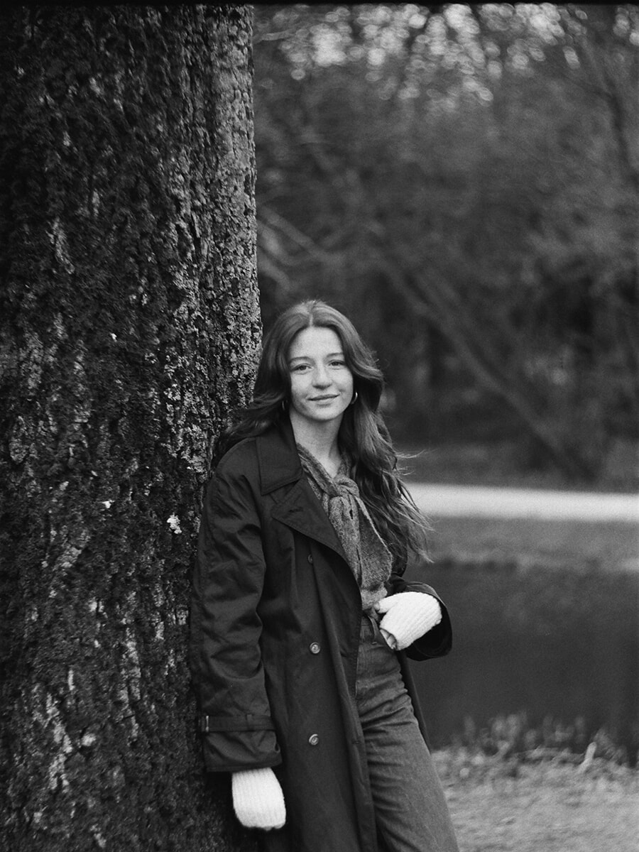 Black-and-white film portrait of a woman in a long coat and scarf leaning against a broad tree trunk